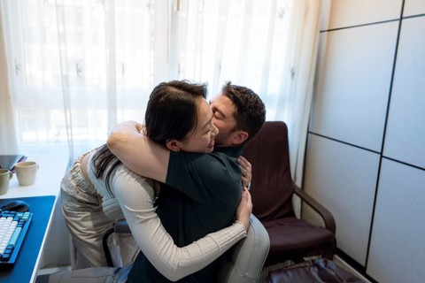 A diverse couple shares a joyful embrace in their cozy home, surrounded by natural light. Their