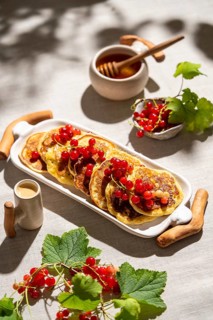 From above, golden pancakes adorned with red currants are beautifully arranged on a white tray.