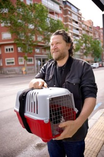 A man carrying a red and white pet carrier with a cat, walking along a city street. The background