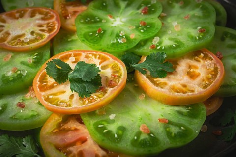 Sliced green tomatoes, on a dark surface, garnished with herbs and salt, top view