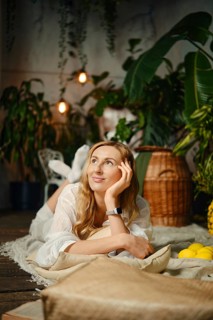 Dreamy woman rests on a soft rug with her chin on her hand, looking up in thought. Green plants