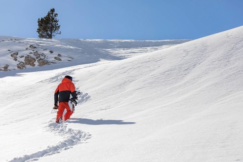 A lone snowboarder in a bright orange outfit climbs a snowy mountain, leaving footprints in the