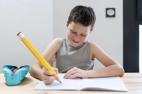 A child, smiling and focused, writes in a notebook using a large yellow pencil. A pencil case is
