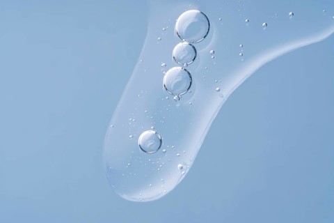 A close-up image of clear, transparent serum drops on a gentle blue background, capturing the light