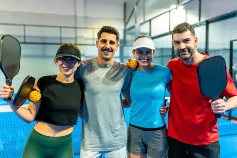 Group of four mixed gender pickleball players embracing and smiling, holding equipment on a blue