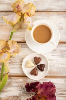 Cup of cioffee with chocolate candies, lilac and purple iris flowers on white wooden background.
