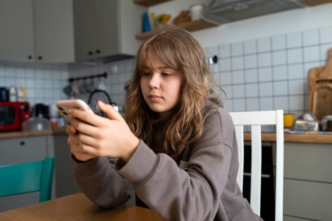 Teen girl sits at a kitchen table focused on her smartphone. Her intent gaze on the screen suggests