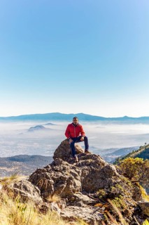 A man in a red jacket sits on a rock, enjoying a panoramic view of the distant mountains and