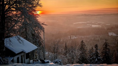 Snowy hut in a wintry mountain landscape at sunset, surrounded by trees and warm light, view from