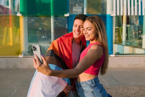 A joyful lesbian couple takes a selfie wrapped in a pride flag. Their smiles and colorful