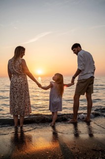 A loving family stands at the edge of the water, holding hands and watching the sun set over the