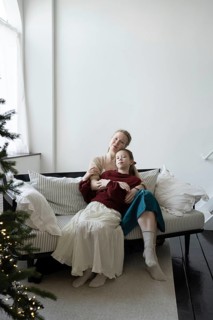 A mother and daughter share a warm embrace on a cozy striped sofa, with a softly lit Christmas tree
