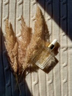 A glass serum cosmetic bottle with golden flakes sits on a textured cream knitted fabric beside