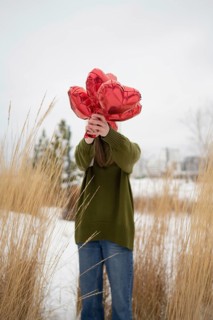 A woman in an olive sweater holds red heart shaped balloons, standing amidst tall grass in a snowy