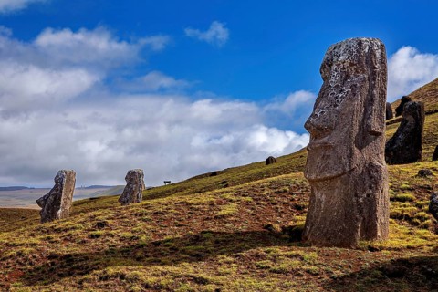 Moai statue against blue sky with clouds on hilly and grassy terrain, The Moai statues on Easter