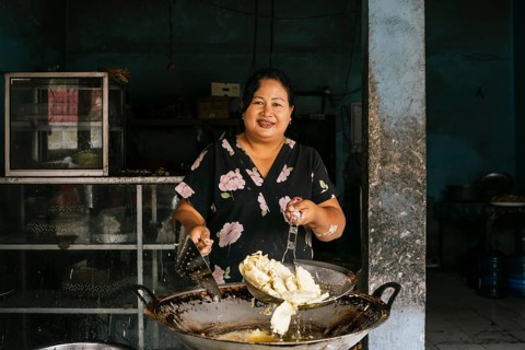 A woman smiles while cooking in a rustic kitchen, holding a frying pan filled with a traditional