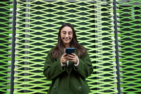A woman wearing a green coat happily uses her smartphone while standing in front of a vibrant green