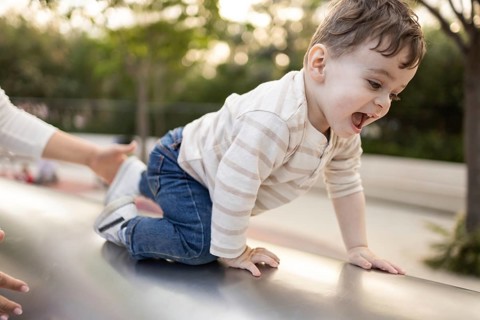 A joyful baby crawls on a slide, with his mother nearby ensuring safety in a vibrant park setting.