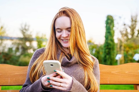 Beautiful red-haired pregnant woman sitting on a park bench, smiling as she looks at her smartphone