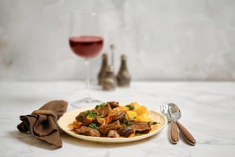 A plate holds pieces of cooked beef and roasted potatoes. A glass of red wine sits nearby. The