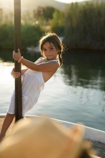 A young girl, dressed in a light summer outfit, playfully stands on a boat in the serene waters of