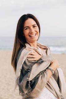 A cheerful woman wrapped in a cozy scarf standing on the beach, smiling sincerely while enjoying