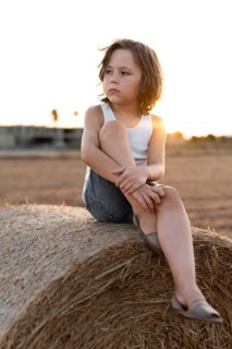 A child in a white tank top and denim shorts sits thoughtfully on a hay bale during sunset. The