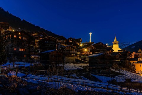 Snow-covered roofs of old farmhouses and barn storage, in the back the illuminated cross and the