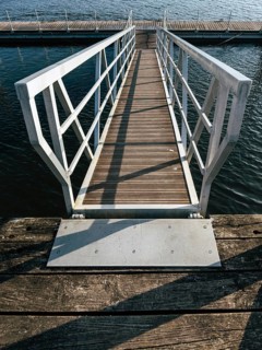 A wooden dock pathway, enclosed by a metal railing, leads to a floating pier over calm water. The