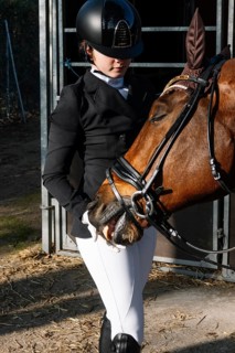A teenage girl in classical dressage attire tenderly interacts with her chestnut horse while