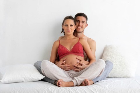 A couple sits on a bed, the pregnant woman in comfortable loungewear, lovingly embraced by her
