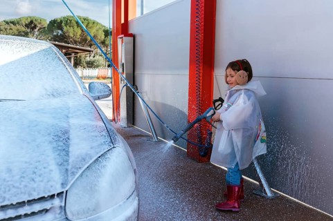 A young child wearing a raincoat and earmuffs joyfully sprays a car with a high pressure hose at a