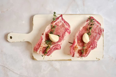 Two raw pork rib chops on a cutting board. Garlic cloves and fresh thyme are placed on top of the
