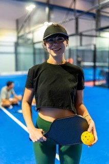 Young woman standing on an indoor blue pickleball court, smiling while holding a paddle and a ball,