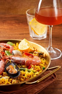 A large dish of seafood paella features shrimp, mussels, and other ingredients. A glass with a