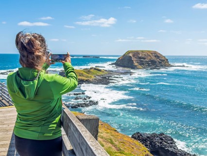 A woman in a green jacket photographs a scenic coastal view with her smartphone in Phillip Island,