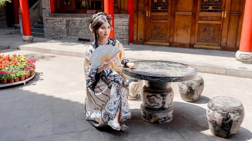 A woman dressed in Qing Dynasty attire is seated outdoors, holding a decorative fan in Pingyao,