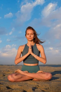 A woman sits in padmasana on the sand at the beach, performing namaste as the sun sets in the