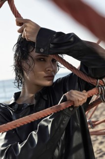 A woman in a black leather jacket holds onto ropes on a sunny beach. Her wet hair and intense gaze