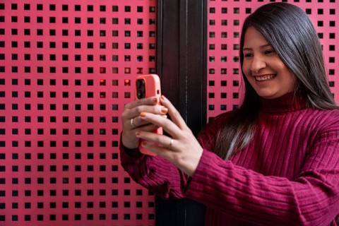 A latin woman in a maroon sweater is smiling as she takes a selfie with her smartphone against a