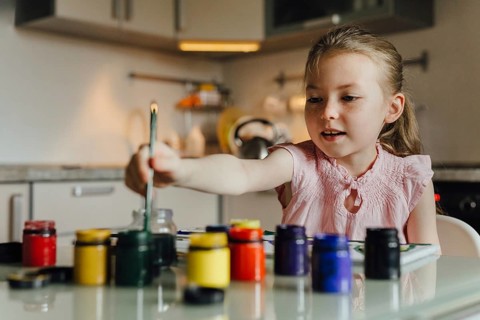 A young girl in a light pink dress joyfully paints with various vibrant colors at home. Art
