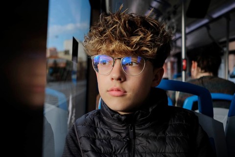 A young man with glasses gazes thoughtfully out the window while riding a city bus. The scene