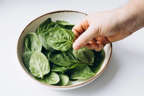 A hand selects fresh spinach leaves from a ceramic bowl. The vibrant green leaves are beautifully