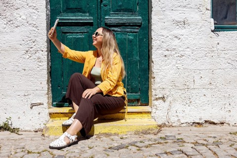 A cheerful woman tourist sits on cobblestone steps, taking a selfie in front of a rustic green