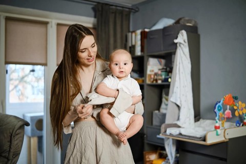A mother with long hair stands in a living room holding her baby. The baby wears a white outfit and