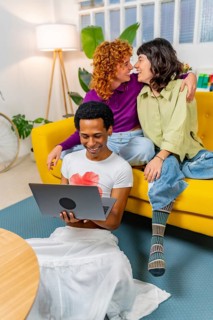 Vertical photo of a Lesbian couple about to kiss next to transgender friend using laptop at home