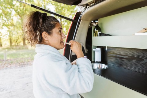 A woman brushes her teeth outside her van, surrounded by the vibrant autumn landscape of New