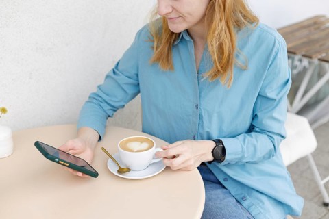 A woman in a blue shirt sits at a cafe table in Warsaw, holding a smartphone and a coffee cup