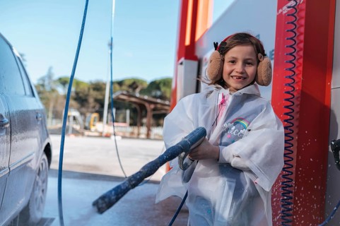 Smiling girl using a high pressure hose at a car wash, wearing a protective raincoat and earmuffs.