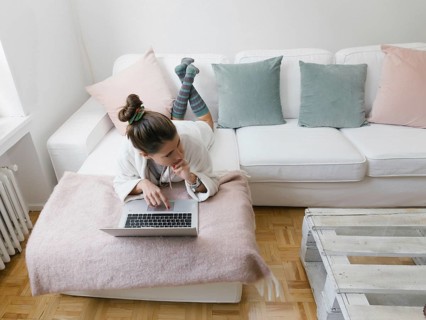 A young woman lies on a white sofa, working on a laptop at home. The living room features soft